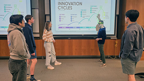Group of students gather around professor as he presents a slide.